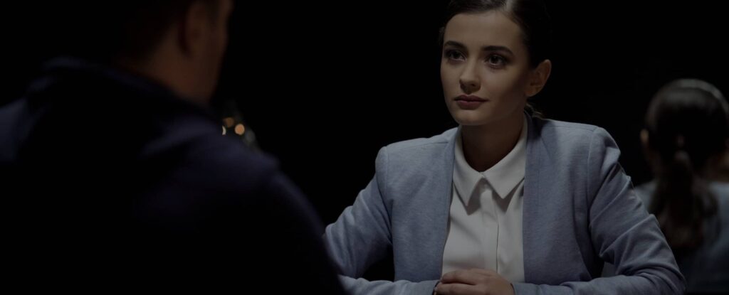 A woman in a light blue blazer sits at a table facing another person in a dimly lit room, appearing to listen attentively.
