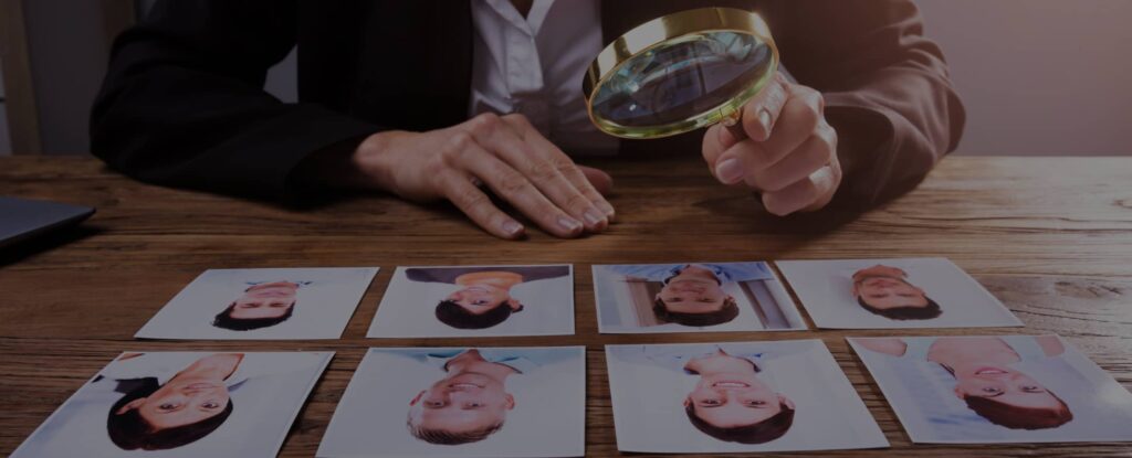 A person examines printed photographs of people on a table using a magnifying glass.