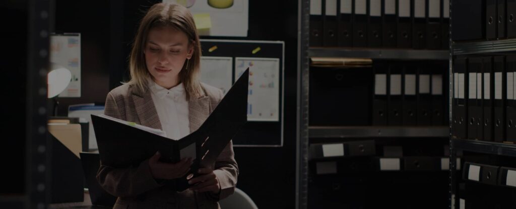 A woman in business attire stands indoors, reading a file folder in front of shelves filled with binders and documents.