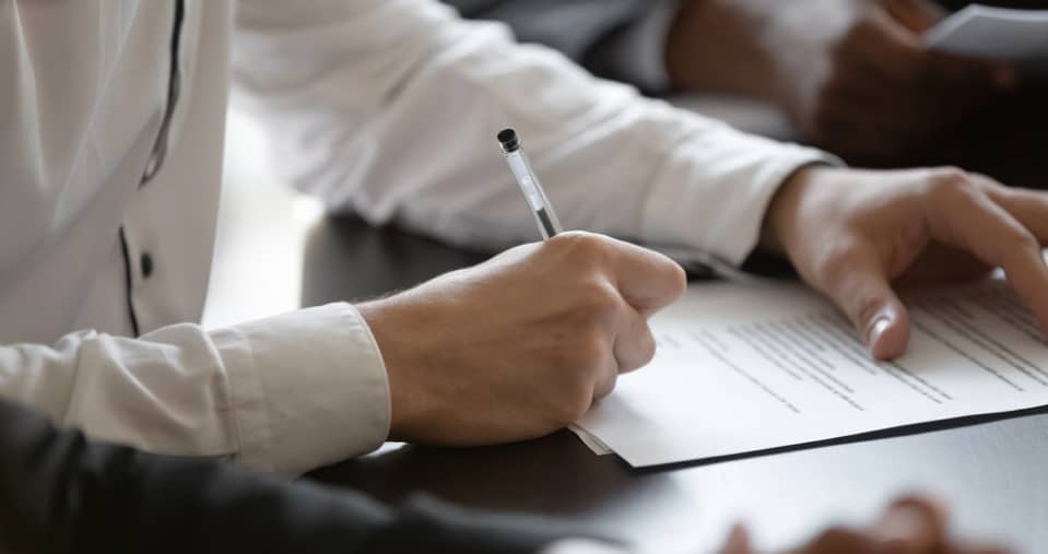 Close-up of a person in a white shirt holding a pen and signing a document on a desk.