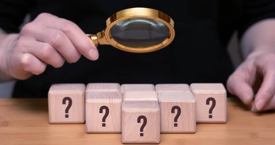 A person holds a magnifying glass over six wooden blocks, each with a question mark on it, placed on a table.