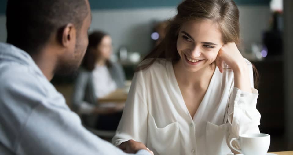 A woman smiles while talking to a man at a table in a café. A cup of coffee is in front of her and another person is blurred in the background.
