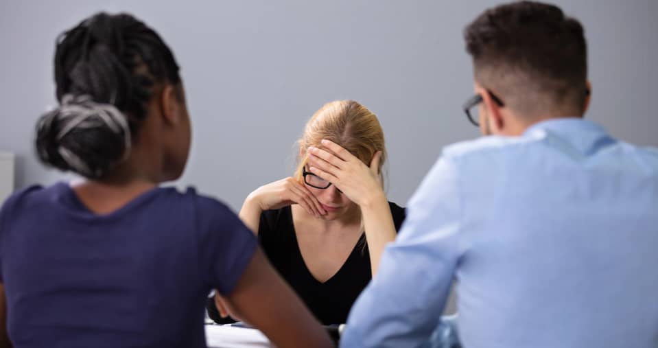 Three people sit at a table; the person in the center holds their head in their hand, appearing stressed, while the other two face them with neutral expressions.